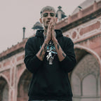 Man in black hoodie with tattoos and sunglasses in front of architectural background